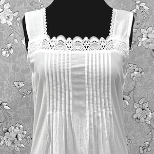 May include: A white sleeveless nightgown featuring a square neckline with intricate lace trim, vertical pintuck pleats on the bodice, and three horizontal stitched lines above a ruffled hemline, displayed on a black mannequin torso.