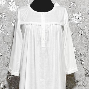 May include: A white long-sleeved nightgown featuring a round neckline with delicate lace trim, a button-down placket on the chest, gathered fabric details, and a ruffled hemline. The garment is displayed on a mannequin against a floral backdrop.