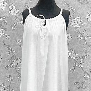May include: A white, floor-length nightgown with a halter neckline and a drawstring closure. The nightgown features a ruffled hem and delicate lace trim. The garment is displayed on a black mannequin against a floral wallpaper backdrop.