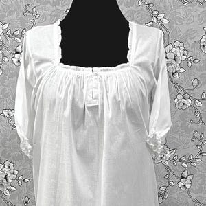 May include: A white, long-sleeved nightgown with a square neckline and gathered bodice. The sleeves are short and finished with a ruffled trim. The nightgown is displayed on a black mannequin against a floral patterned background.