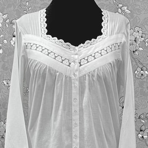 May include: A long, white nightgown with long sleeves and a button-down front. The nightgown features delicate lace trim at the neckline, cuffs, and hem. The fabric appears lightweight and flowing, with subtle pleating details.