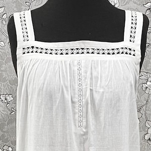 May include: A white nightgown with a square neckline and delicate lace trim. The sleeveless gown has a long, flowing design and a subtle vertical seam detail down the front. The fabric appears to be lightweight and breathable.