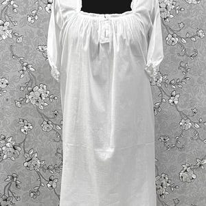 May include: A white, knee-length nightgown with short sleeves and a square neckline. The neckline and sleeve cuffs have a ruffled trim. The fabric appears to be lightweight and slightly sheer. The nightgown is displayed on a black mannequin against a floral patterned background.
