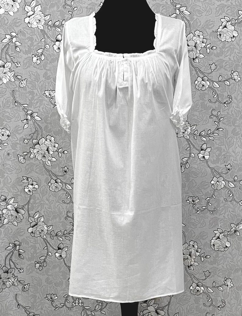 May include: A white, knee-length nightgown with short sleeves and a square neckline. The neckline and sleeve cuffs have a ruffled trim. The fabric appears to be lightweight and slightly sheer. The nightgown is displayed on a black mannequin against a floral patterned background.