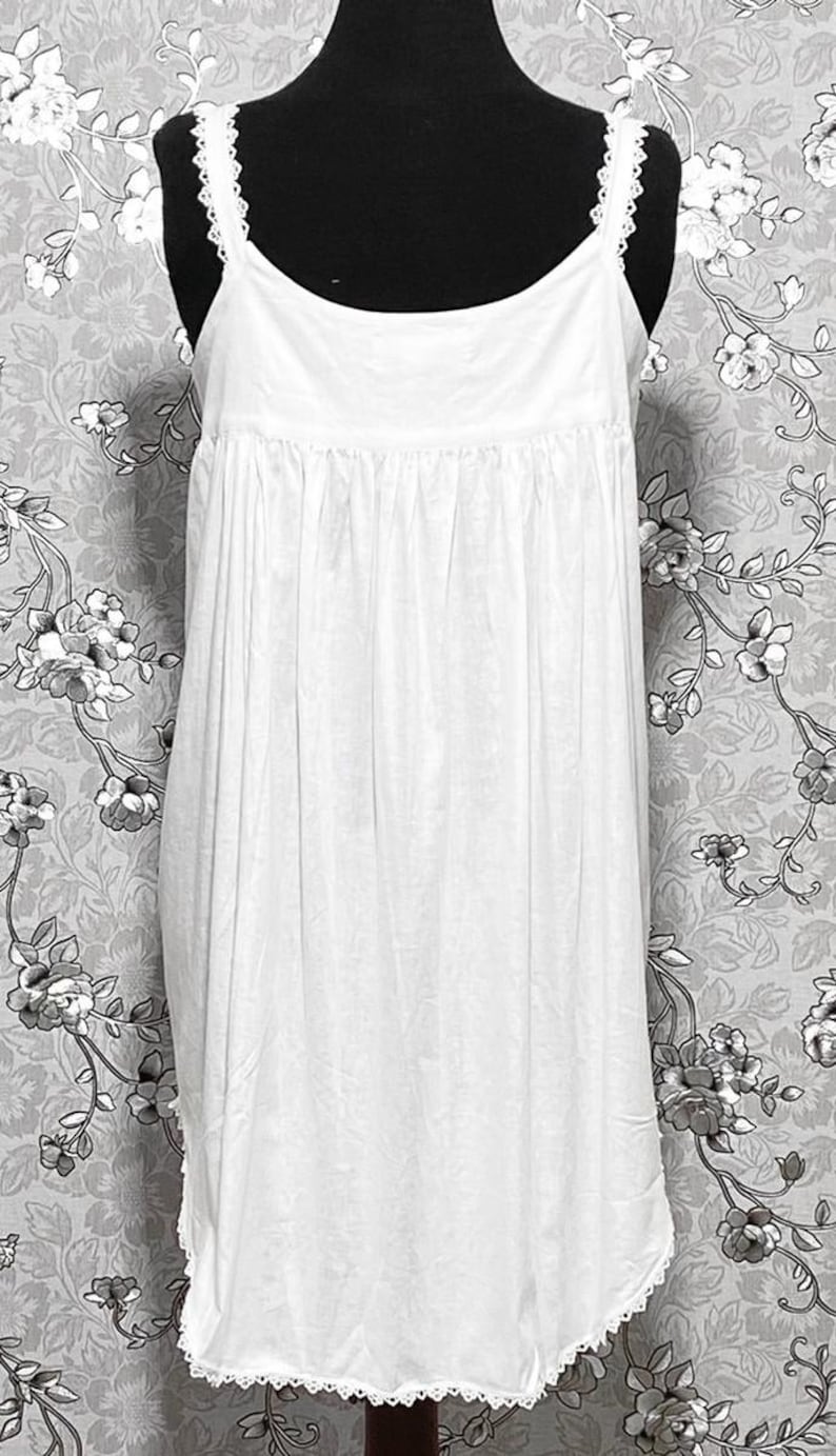 Peut inclure: Une chemise de nuit en coton blanc avec des bordures en dentelle. La chemise de nuit a un col rond et des bretelles fines.