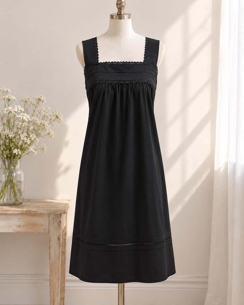 May include: A black knee-length dress featuring a square neckline, wide shoulder straps, and delicate scalloped lace trim along the bodice. The garment includes a gathered waistline and a horizontal lace insert detail near the hemline.