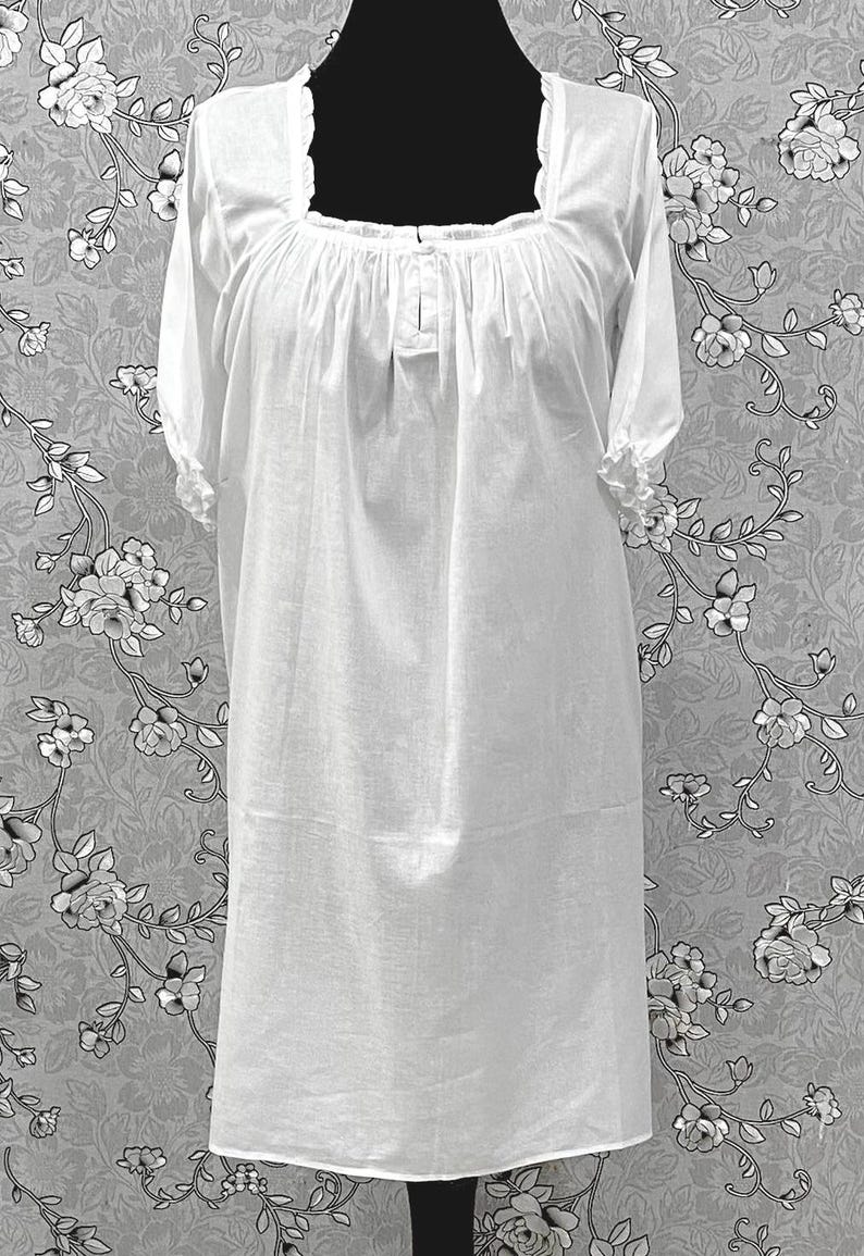 May include: A white, long-sleeved nightgown with a square neckline and gathered bodice. The sleeves are short and finished with a ruffled trim. The nightgown is displayed on a black mannequin against a floral patterned background.