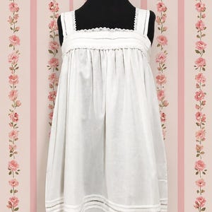May include: A white sleeveless dress featuring a square neckline with delicate lace trim, wide shoulder straps, and a gathered bodice. The hem is detailed with horizontal bands of openwork embroidery and stitched fabric panels.