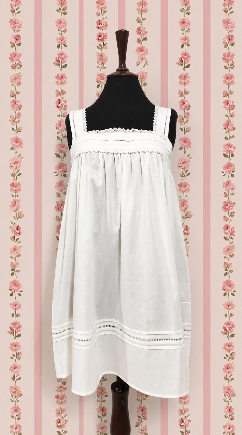 May include: A white sleeveless dress featuring a square neckline with delicate lace trim, wide shoulder straps, and a gathered bodice. The hem is detailed with horizontal bands of openwork embroidery and stitched fabric panels.