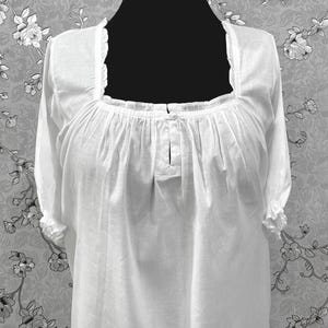 May include: A white, long-sleeved blouse with a square neckline and ruffled trim. The blouse features gathered fabric details at the neckline and sleeves. The garment is displayed on a black mannequin.