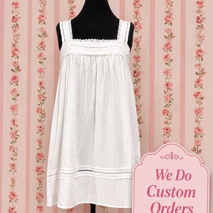 May include: A white sleeveless dress featuring a square neckline with delicate lace trim, a gathered bodice, and horizontal pintuck detailing near the hem. The dress is displayed on a black mannequin against a pink and white floral striped background.