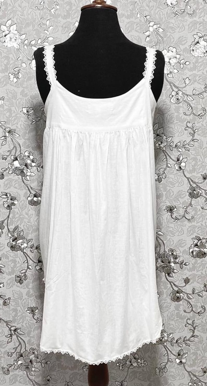 Peut inclure: Robe de nuit blanche en coton avec une bordure en dentelle autour de l'encolure et de l'ourlet. La robe a un corsage pliss&eacute; et une jupe ample.