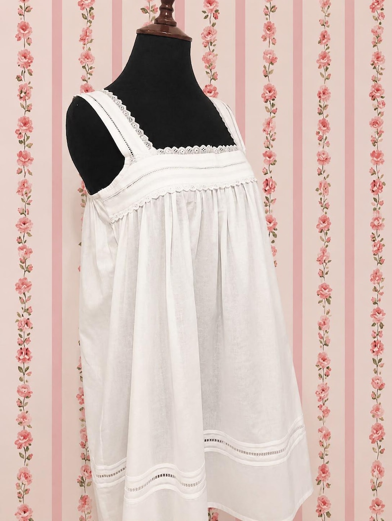 May include: A white sleeveless nightgown featuring a square neckline with delicate lace trim, wide shoulder straps, and vertical pleats across the chest. The hem is detailed with horizontal bands of openwork embroidery and matching lace trim.