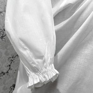 May include: Close-up of a white long-sleeved garment. The sleeve features a gathered cuff with a ruffled edge. The fabric appears to be lightweight and slightly textured, with soft folds and shadows.