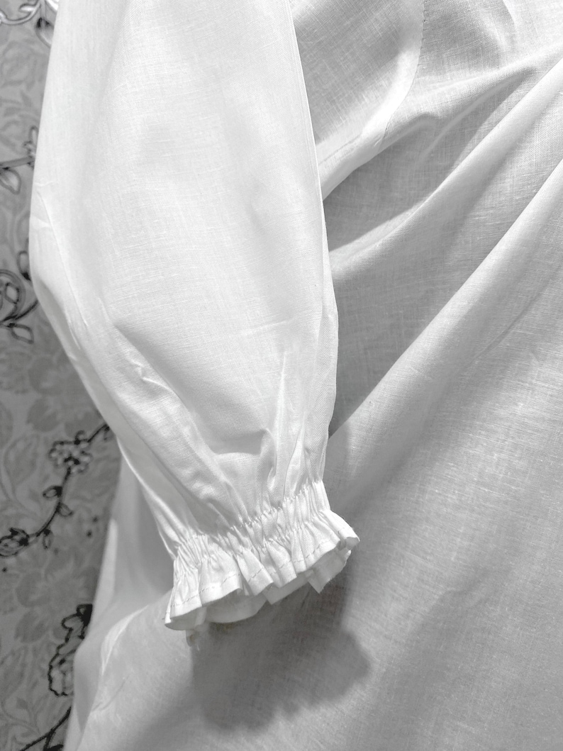 May include: Close-up of a white long-sleeved garment. The sleeve features a gathered cuff with a ruffled edge. The fabric appears to be lightweight and slightly textured, with soft folds and shadows.