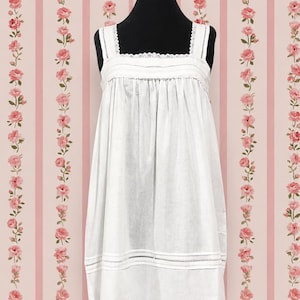 May include: A white sleeveless chemise featuring a square neckline with delicate lace trim, gathered fabric at the bodice, and horizontal pintuck detailing along the lower hem. The garment is displayed on a black mannequin torso.