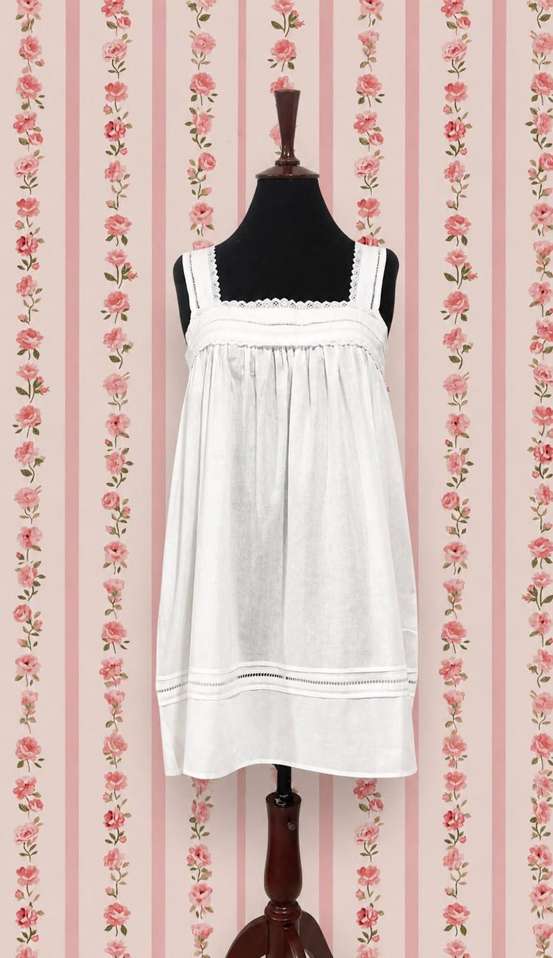 May include: A white sleeveless chemise featuring a square neckline with delicate lace trim, gathered fabric at the bodice, and horizontal pintuck detailing along the lower hem. The garment is displayed on a black mannequin torso.