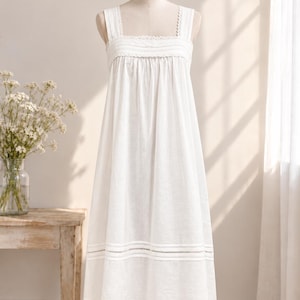 May include: A white sleeveless dress featuring a square neckline with delicate lace trim, a gathered bodice, and a wide horizontal band of openwork embroidery detail near the hemline. The dress has a loose, flowing silhouette and wide shoulder straps.