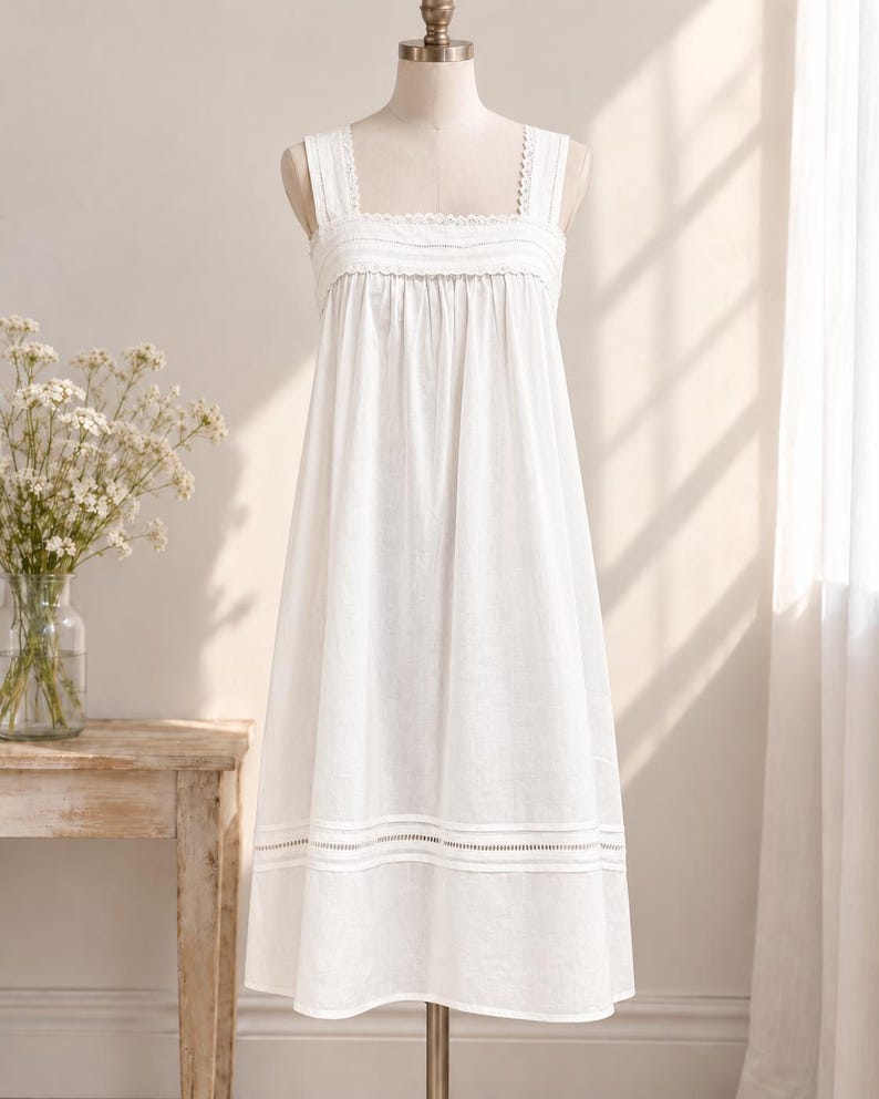 May include: A white sleeveless dress featuring a square neckline with delicate lace trim, a gathered bodice, and a wide horizontal band of openwork embroidery detail near the hemline. The dress has a loose, flowing silhouette and wide shoulder straps.