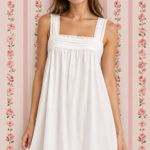 May include: A white sleeveless dress featuring a square neckline with delicate lace trim, a gathered bodice, and a horizontal lace insert detail near the hemline. The garment has a simple, loose-fitting silhouette and a clean, solid white color.