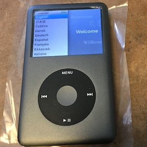 May include: A silver iPod with a black control wheel and screen displaying a welcome message in multiple languages. The screen shows a list of languages including English, Japanese, and Spanish. The device is in a clear plastic wrapper.