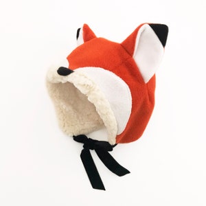 Peut inclure: Un chapeau renard orange avec des détails blancs et noirs. Le chapeau a une doublure en fausse fourrure douce et des liens en ruban noir. Le chapeau a des oreilles pointues et un visage de renard.