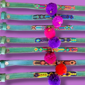 May include: Seven teal leather collars numbered 64 through 70, each featuring a central woven textile strip with geometric patterns in various colors and a single hanging pom-pom in purple, pink, or orange attached to the center.