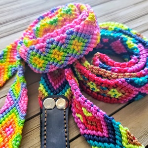 May include: A long, multi-colored woven strap featuring a repeating diamond pattern in vibrant shades of neon pink, yellow, green, blue, and orange. The strap is attached to a black leather end piece secured with two silver-toned metal rivets.
