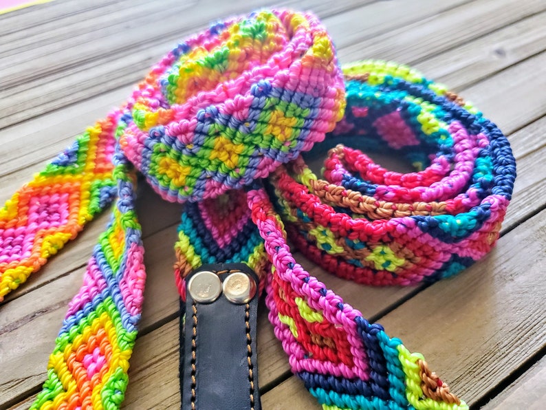 May include: A long, multi-colored woven strap featuring a repeating diamond pattern in vibrant shades of neon pink, yellow, green, blue, and orange. The strap is attached to a black leather end piece secured with two silver-toned metal rivets.