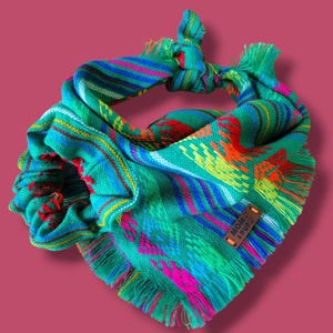 May include: A green, blue, pink, and yellow patterned bandana with fringe. The bandana has a brown leather tag with the text "MOM & PUP" on it.