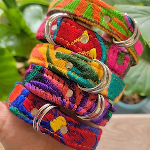 Puede incluir: Una pila de cinco collares de tela con bordados florales vibrantes y multicolores en tonos naranja, rosa, verde, amarillo, morado y rojo. Cada collar incluye anillas en D de metal plateado y remaches circulares para su sujeción.