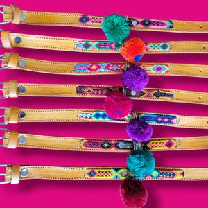 May include: Seven tan leather belts labeled 8 through 14, each featuring a central woven textile strip with colorful geometric patterns. Each belt includes a silver metal buckle and a single hanging yarn pom-pom in various bright colors.