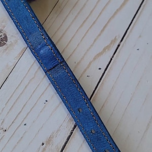 May include: A blue leather strap featuring orange contrast stitching along the edges, a silver metal eyelet, and a fuzzy purple pom-pom attached to the end. The strap has a series of evenly spaced holes for adjustment and a textured surface finish.