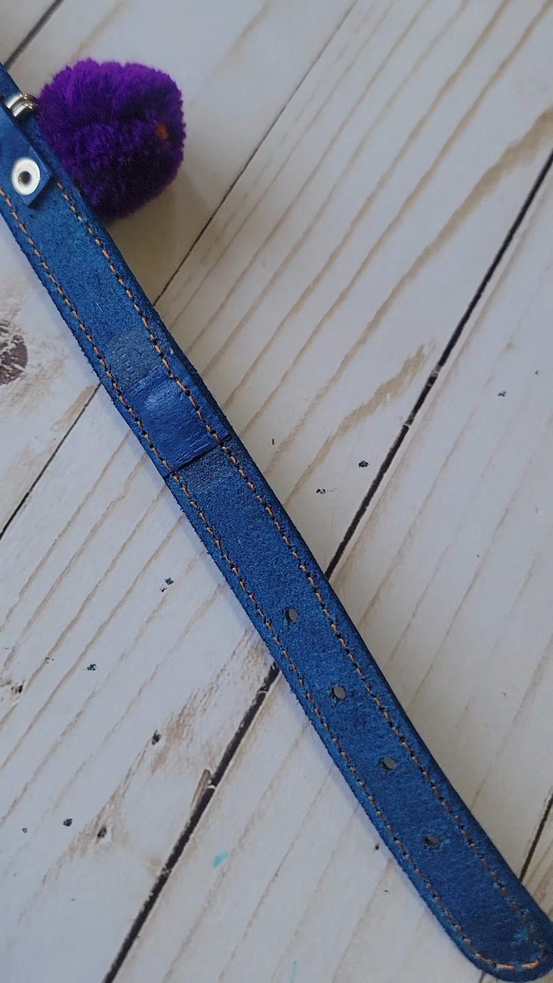 May include: A blue leather strap featuring orange contrast stitching along the edges, a silver metal eyelet, and a fuzzy purple pom-pom attached to the end. The strap has a series of evenly spaced holes for adjustment and a textured surface finish.