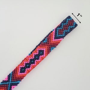 May include: A woven macrame bracelet featuring a geometric pattern in navy blue, dark red, bright pink, and light blue threads. The band has a width of 1 inch as indicated by the measurement text and arrow graphic visible at the top edge.