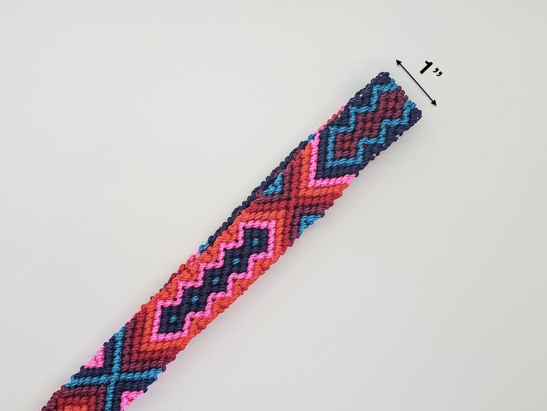 May include: A woven macrame bracelet featuring a geometric pattern in navy blue, dark red, bright pink, and light blue threads. The band has a width of 1 inch as indicated by the measurement text and arrow graphic visible at the top edge.