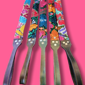May include: Five long, narrow straps featuring vibrant, multi-colored floral embroidery on fabric. Each strap is attached to a leather end piece with three silver-toned metal rivets. The leather ends vary in shades of brown, green, and black.