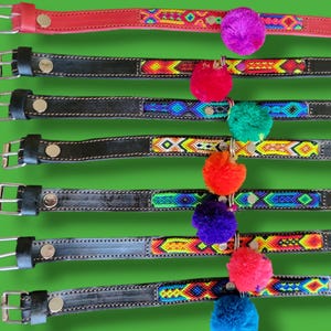 May include: A collection of seven leather belts numbered 36 through 42, featuring silver-toned metal buckles and colorful woven fabric inlays. Each belt is adorned with a single vibrant pom-pom in shades of purple, red, green, orange, or blue.