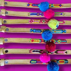 May include: Seven tan leather collars labeled 50 through 56, each featuring a central woven textile strip with geometric patterns in vibrant colors and a single large, fluffy pom-pom in blue, pink, yellow, or red attached to the center.