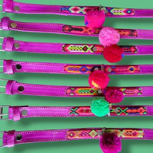 May include: Seven bright pink leather collars numbered one through seven, each featuring a unique woven fabric inlay with geometric patterns and a single colorful yarn pom-pom attached to a metal ring near the center of the strap.