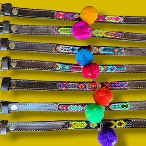 May include: Seven dark brown leather collars numbered 15 through 21, each featuring a central woven fabric strip with geometric patterns in vibrant colors and a single large, round, fuzzy pom-pom attached to the center of each collar.