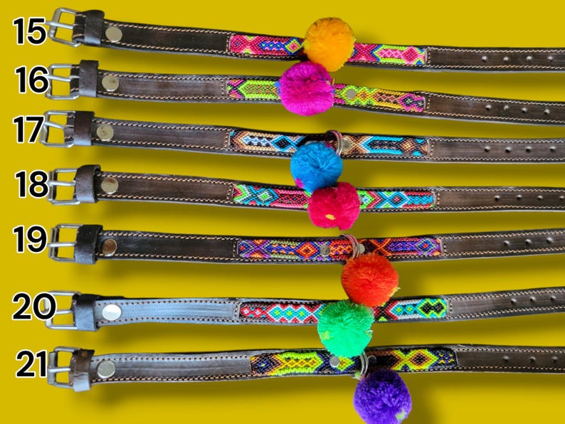 May include: Seven dark brown leather collars numbered 15 through 21, each featuring a central woven fabric strip with geometric patterns in vibrant colors and a single large, round, fuzzy pom-pom attached to the center of each collar.
