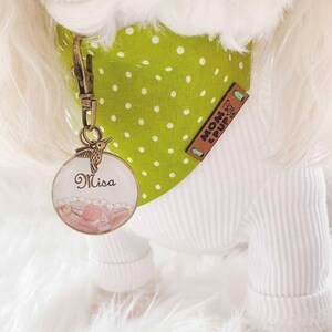 May include: A white dog wearing a green and white polka dot bandana with a brown leather tag that says "Mom & Pup Co." The dog is also wearing a personalized name tag with a hummingbird design and the name "Misa" written on it.