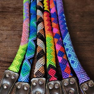 May include: Six braided cord leashes featuring intricate geometric patterns in vibrant colors including neon green, blue, brown, black, orange, pink, and purple. Each cord is attached to a dark brown leather end piece secured with three silver rivets.