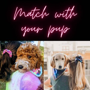 May include: Two women are holding their dogs. Both women are wearing scrunchies in their hair and their dogs are wearing bandanas. The text "Match with your pup" is displayed in pink neon letters.