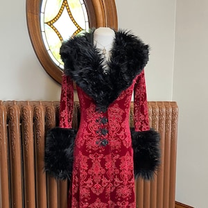 Op de afbeelding: Een lange rode fluwelen robe met een zwarte nep bontkraag en manchetten. De robe heeft een damastpatroon en een knoopsluiting.