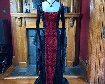 May include: A long black velvet gown with a red velvet panel down the front. The gown has long sleeves with lace detailing and a square neckline. The gown is being displayed on a mannequin in a room with a fireplace.