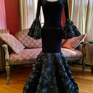 May include: A black velvet gown with long sleeves and a fitted bodice. The skirt is made of black satin and features a large, dramatic rose design.