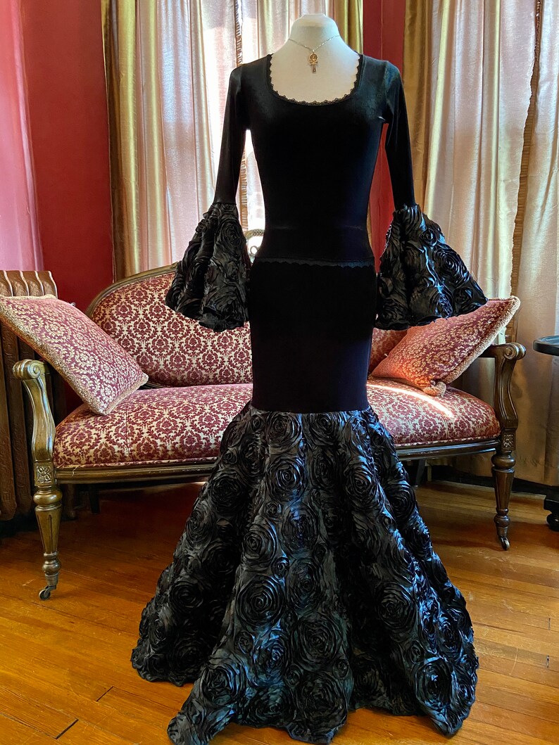 May include: A black velvet gown with long sleeves and a fitted bodice. The skirt is made of black satin and features a large, dramatic rose design.