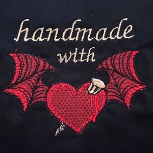 May include: Embroidered design with the text "handmade with" above a red heart with wings. The heart has a small white object on top and a red drip coming from it.